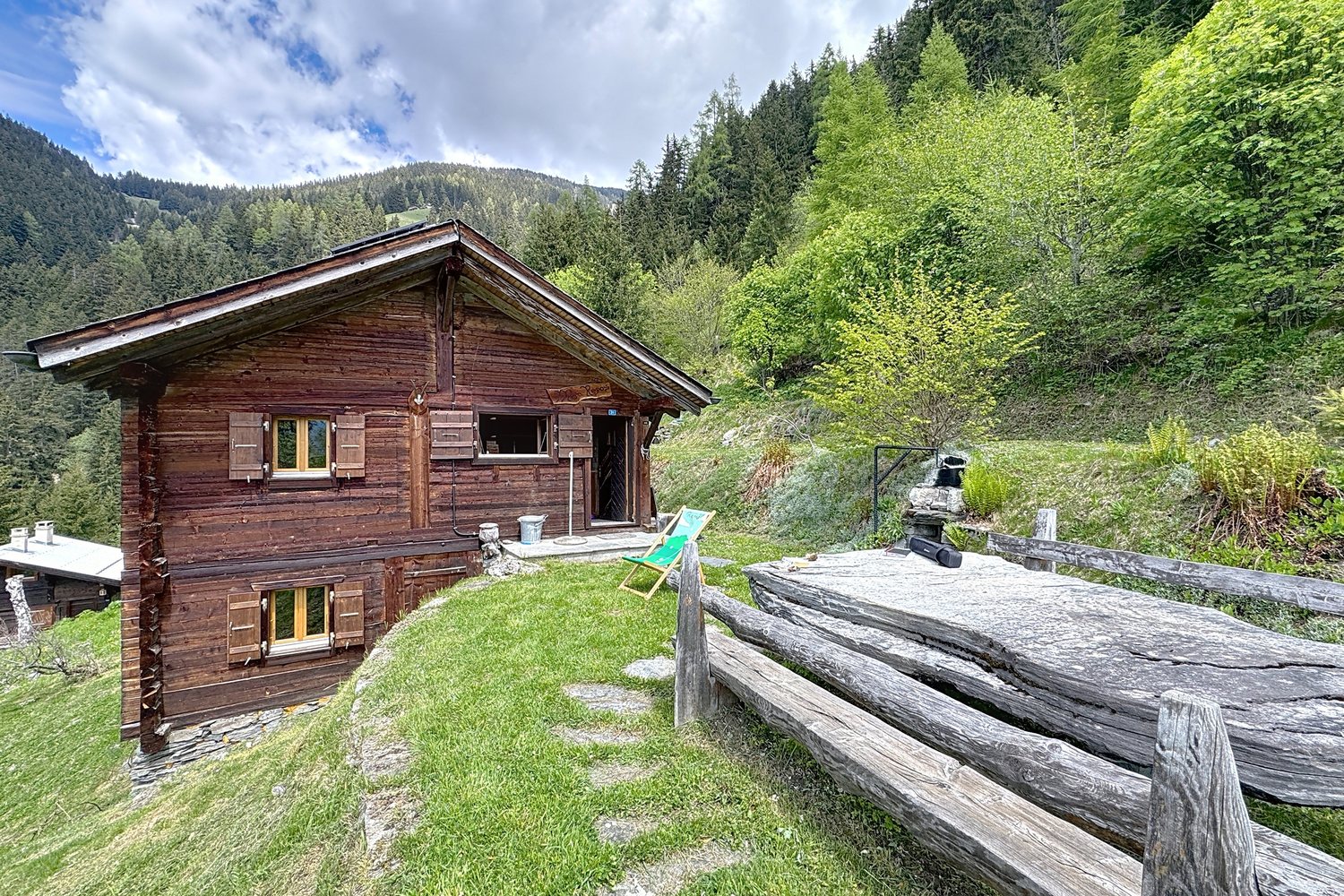 Le chalet vu du chemin, banc de bois au premier plan, sentier dallé qui monte vers la porte, forêt derrière.
