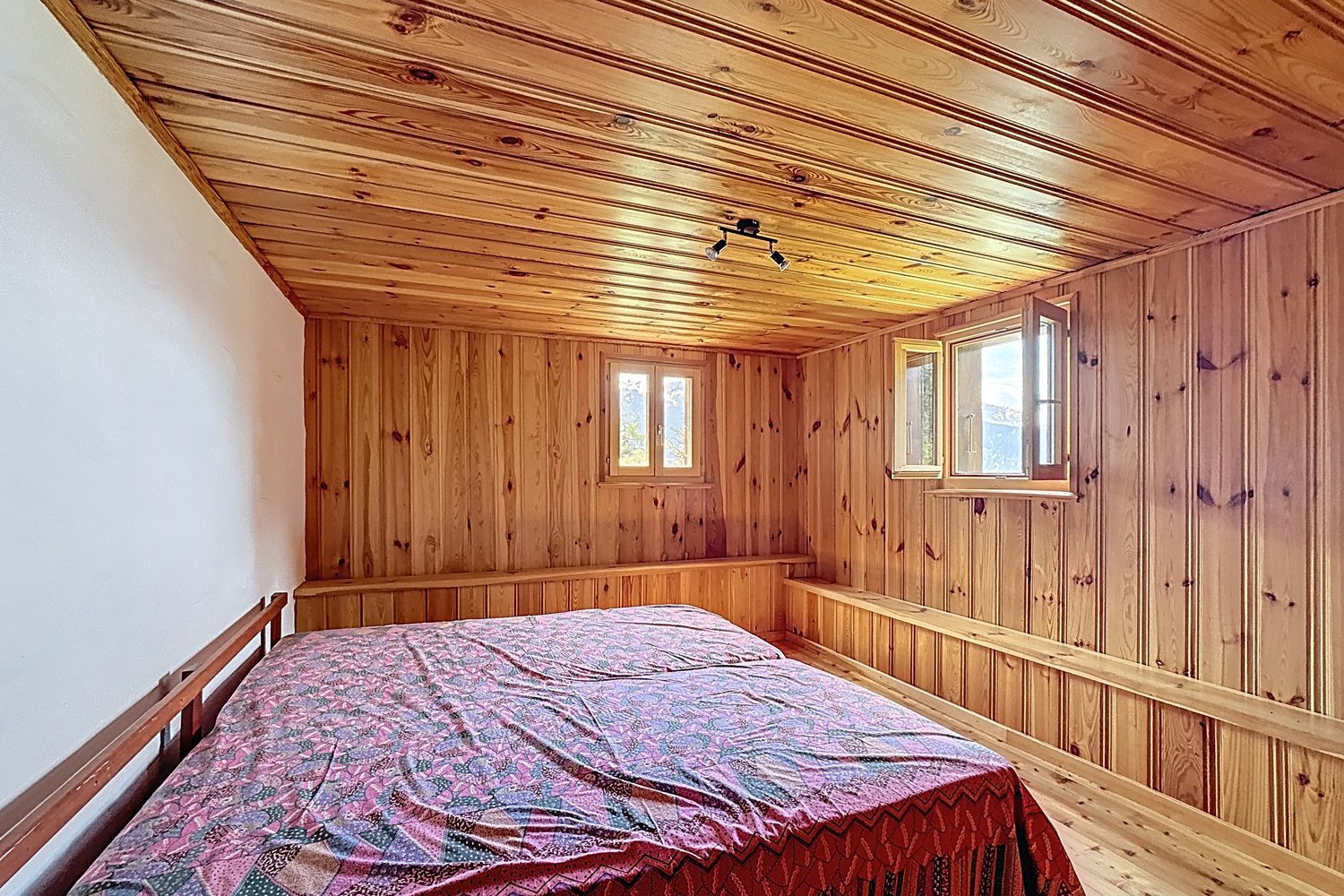 Chambre lambrissée du chalet : lit double avec couvre-lit rouge sous la fenêtre, plafond en pente.