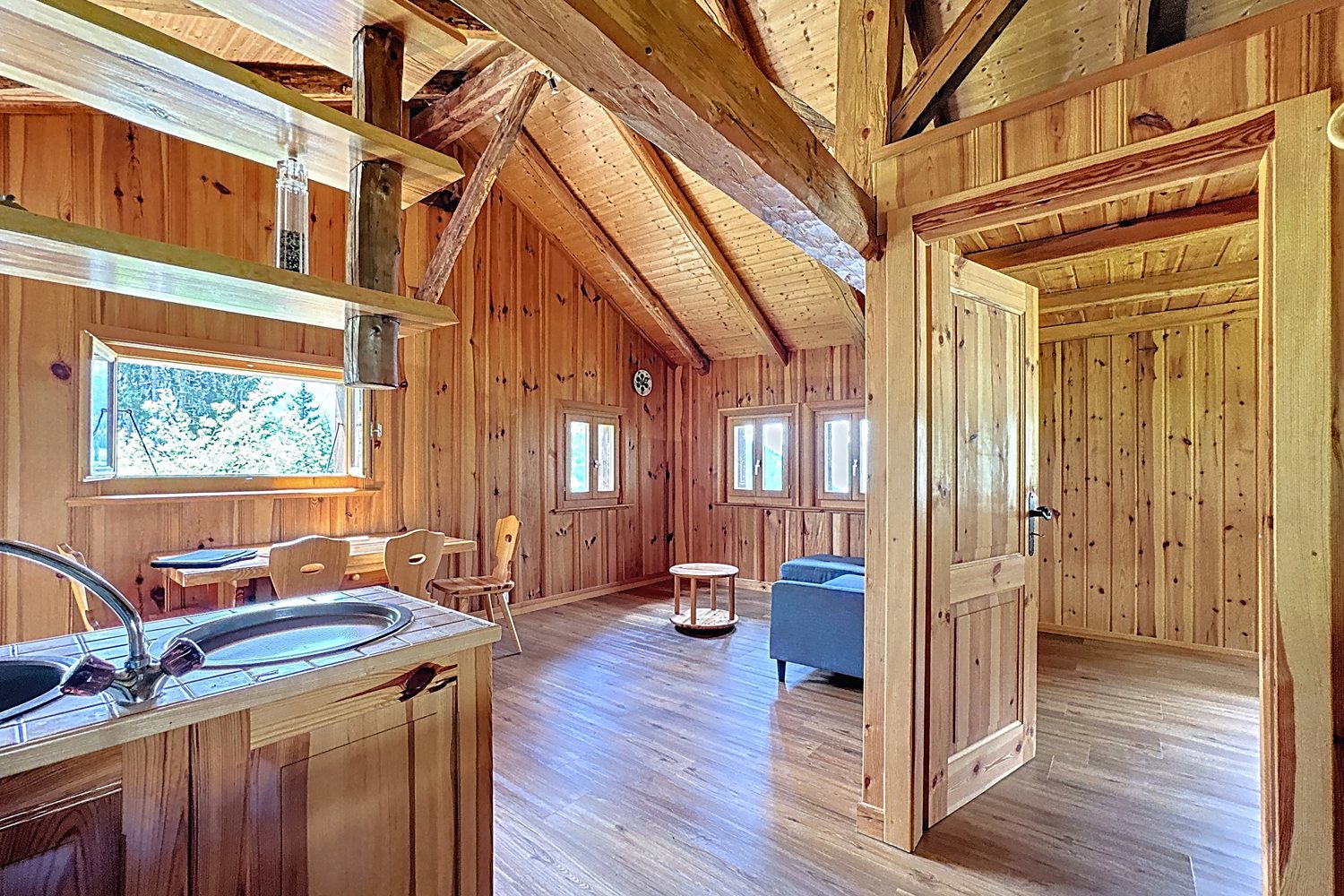 Coin repas du chalet : table et chaises en bois sous la fenêtre, évier au premier plan, porte vers la seconde chambre.