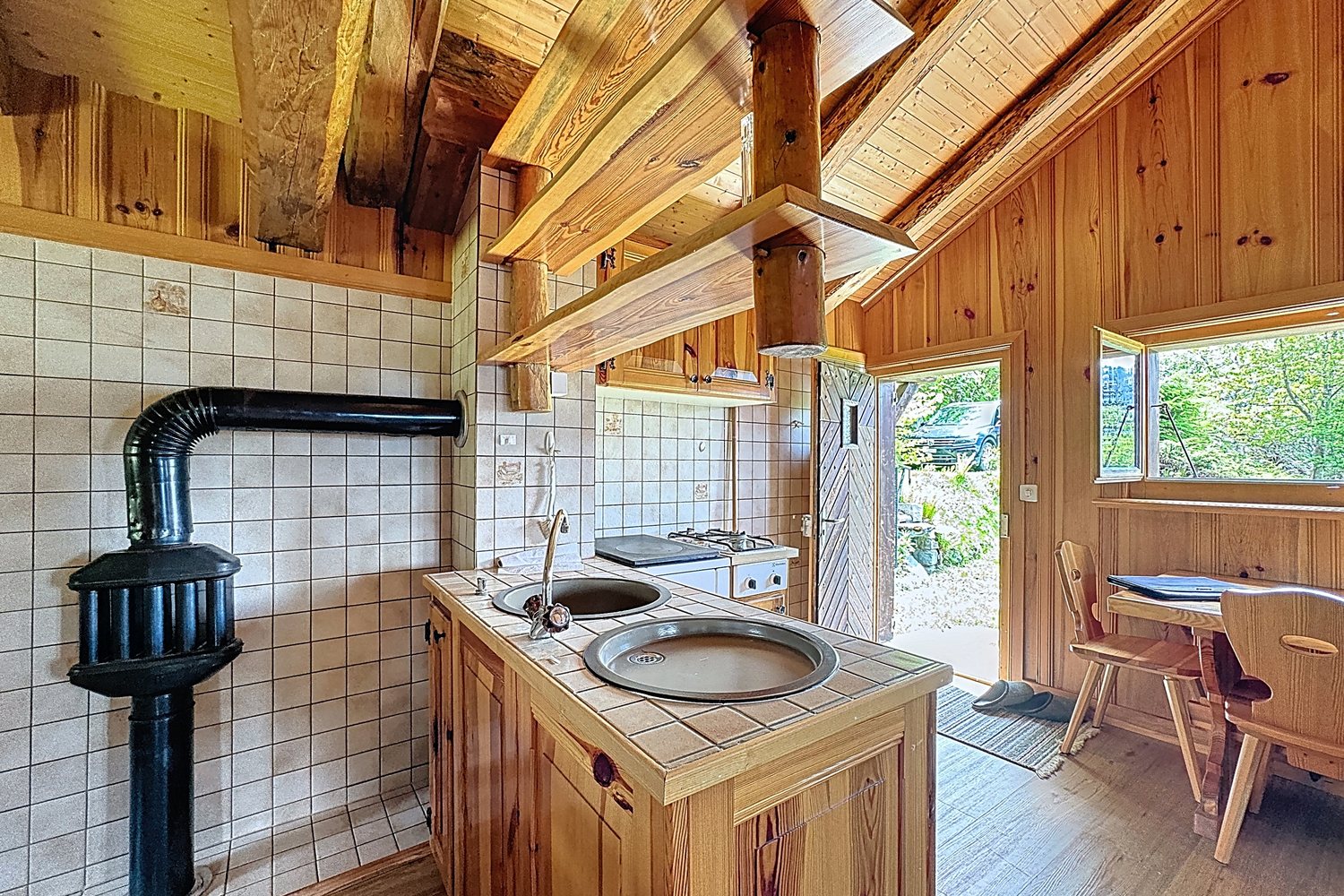 Cuisinière à bois noire contre un mur carrelé blanc, évier, et porte ouverte sur le pré et les arbres.