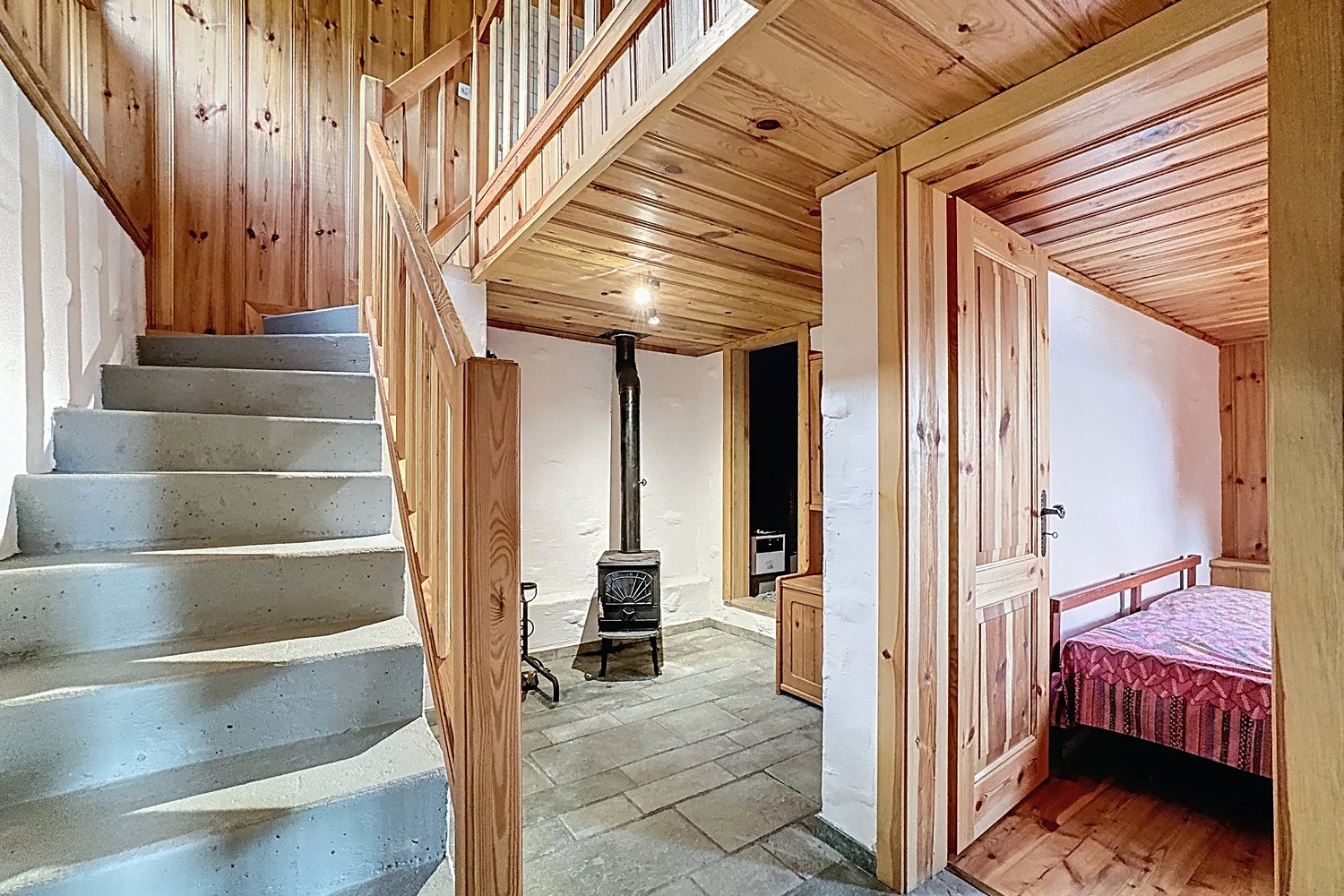 Escalier en béton lissé du chalet, poêle à bois au pied des marches, porte ouverte sur la chambre.