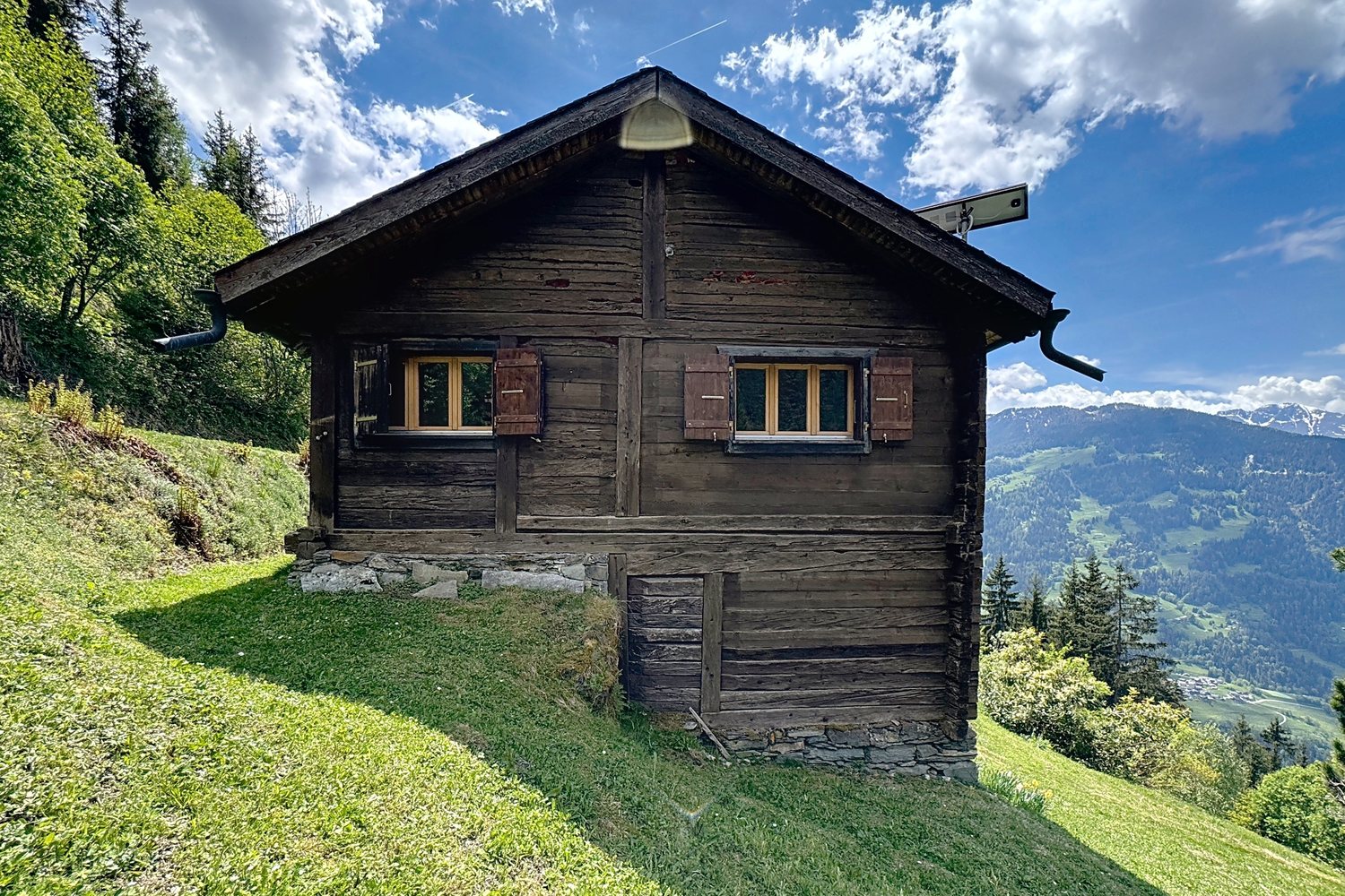 Façade du chalet en madriers sombres sur socle de pierre, volets ouverts, pré tondu devant et la vallée de Bagnes sur le côté.