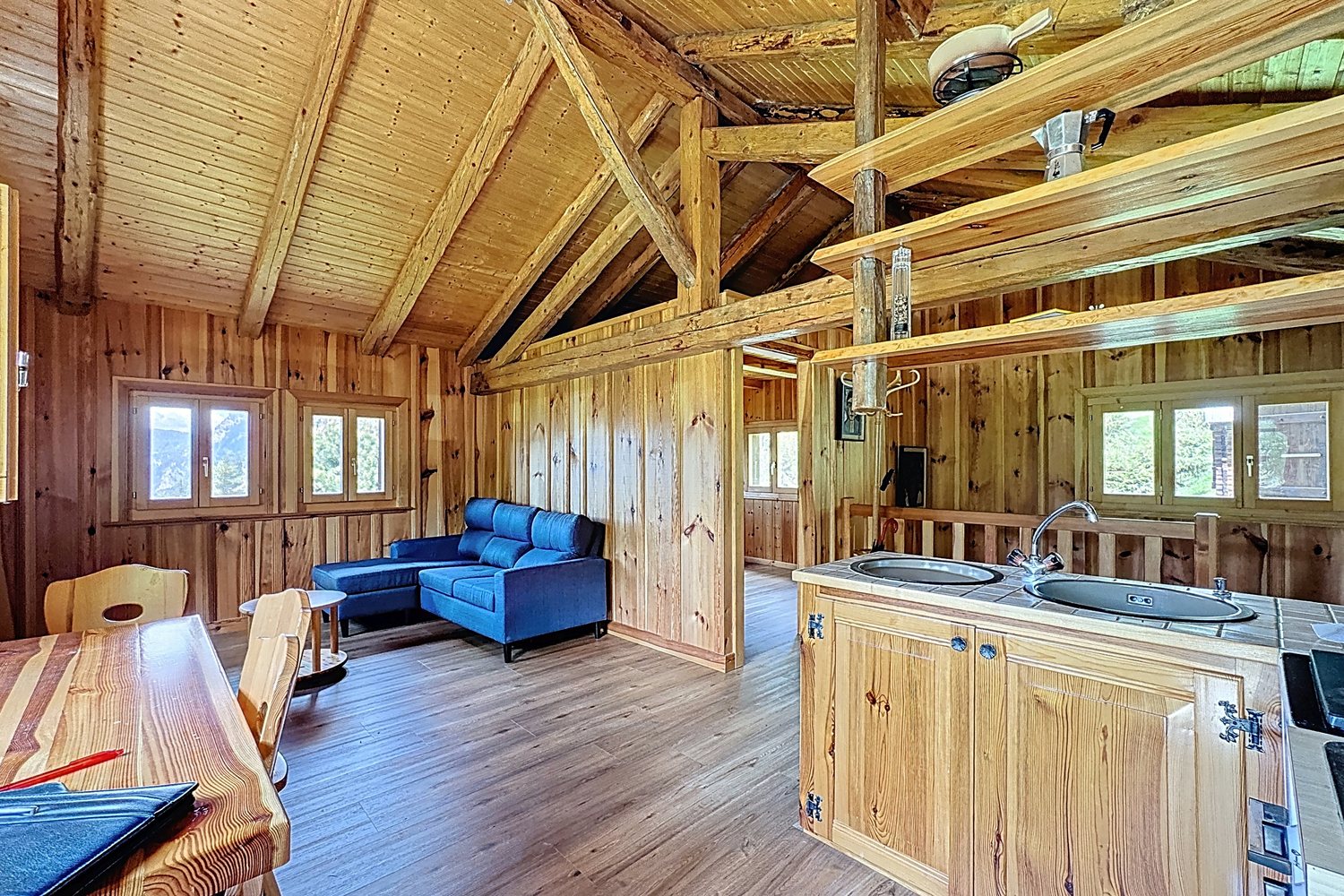 Pièce de vie du chalet : charpente apparente, lambris clair, parquet, canapé bleu sous les fenêtres, îlot de cuisine en bois.
