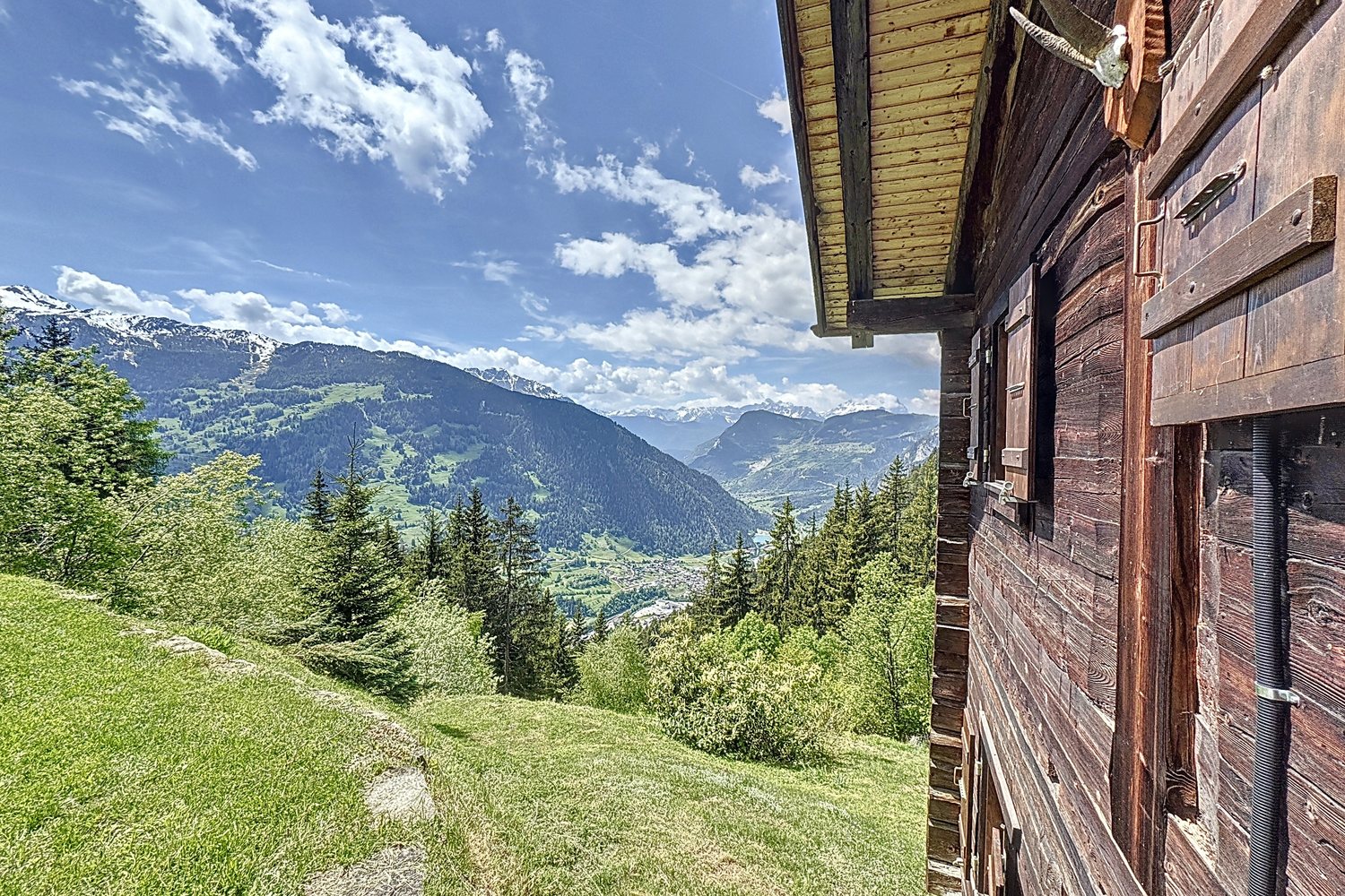 Vue depuis l'angle du chalet : les madriers au premier plan, la vallée de Bagnes, les forêts et les sommets enneigés au loin.